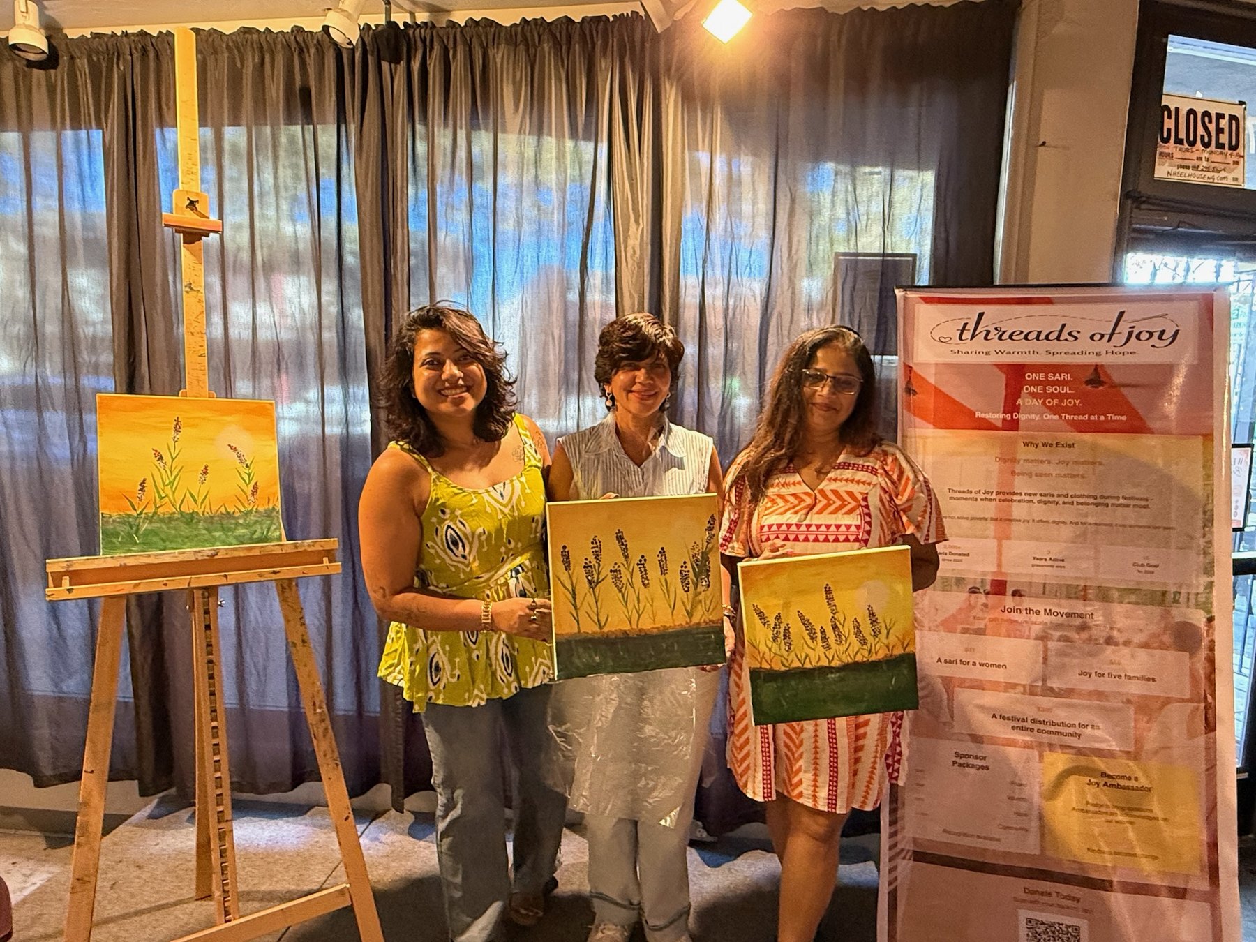 Threads of Joy organizers stand near the event banner with completed paintings.